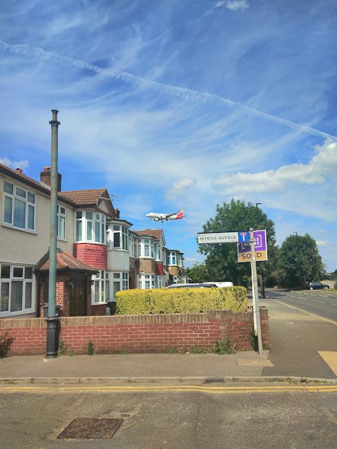 A clear daytime scene of residential houses along Ewell Road, with a red-brick and white-facade terraced property featuring multiple front-facing bay windows and a small front garden bordered by a low brick wall. To the right, a large directional road sign indicates nearby streets, including Myrtle Avenue and Tolworth Tower, while a white commercial van is parked on the pavement. An airplane is captured mid-flight in the bright blue sky, which is partly cloudy with high-altitude contrails. A street lamp with a black post stands near the sidewalk, and the road surface shows double yellow lines, indicating parking restrictions. The environment suggests a suburban area, possibly during a house removal or relocation process, with the presence of the van and signs of potential moving activity. Man with Van Tolworth's services are implied through the context of moving logistics and furniture transport, highlighting the typical surroundings for home relocation in the KT5 area.