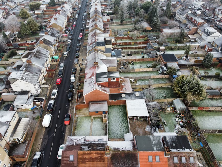 An aerial view of a residential street during daytime, showing lined houses with pitched roofs, some with snow on top. The street has parked cars on both sides, and a van is parked adjacent to a house near the bottom of the image. Several fences and gardens are visible behind the houses, with some gardens containing outdoor furniture, sheds, and small trees. The scene captures a typical neighbourhood setup suitable for house removals and home relocation logistics, with clear access to properties and a road suitable for vehicle loading and unloading. This environment reflects the typical setting for furniture transport, packing, and loading processes undertaken by Man with Van Tolworth during house moves in the KT5 postcode area.