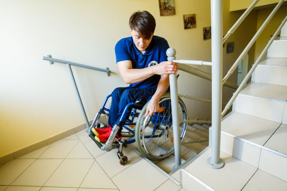 A person in a blue shirt, seated in a wheelchair with red and yellow shoes, is holding and stabilising a white handrail on the wall at the top of a staircase in an indoor setting. The staircase has beige steps, and the handrail is attached to a pale yellow wall, with a metal guardrail visible along the staircase. The floor is tiled with light-colored ceramic tiles, and a small section of a wall with some framed pictures can be seen. The scene appears to depict a moving or packing process related to house removals, with the individual possibly assisting or preparing for furniture transport or home relocation, facilitated by Man with Van Tolworth. The environment is well-lit, and the overall setting suggests accessibility considerations during a flat-to-flat move within a residential property.