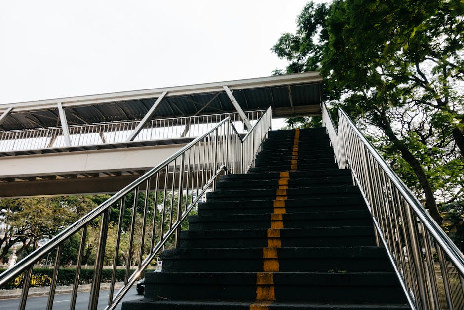 A set of outdoor stairs with black steps and a yellow safety line running down the middle, bordered by silver metal handrails on both sides, leading up to an elevated pedestrian bridge with a metal roof and white railings. The stairs are situated outside a residential or commercial building, surrounded by green trees and clear sky. The image captures the logistical challenge of navigating stair access during a house or flat move, as often managed by professional removal companies like Man with Van Tolworth, highlighting the importance of careful planning during home relocation or furniture transport projects.