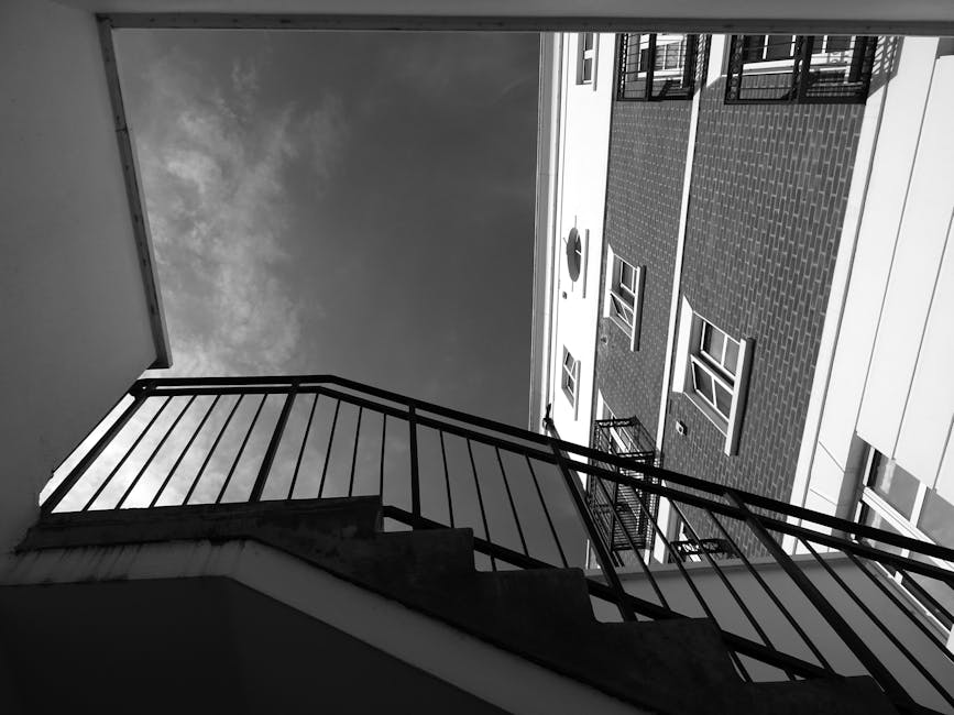 A person in a blue shirt, seated in a wheelchair with red and yellow shoes, is holding and stabilising a white handrail on the wall at the top of a staircase in an indoor setting. The staircase has beige steps, and the handrail is attached to a pale yellow wall, with a metal guardrail visible along the staircase. The floor is tiled with light-colored ceramic tiles, and a small section of a wall with some framed pictures can be seen. The scene appears to depict a moving or packing process related to house removals, with the individual possibly assisting or preparing for furniture transport or home relocation, facilitated by Man with Van Tolworth. The environment is well-lit, and the overall setting suggests accessibility considerations during a flat-to-flat move within a residential property.