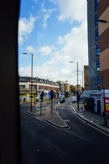 A view through a vehicle window showing a street scene on a bright day with partly cloudy skies. The street is lined with multi-storey buildings, including a shopping arcade with signage and shopfronts. In the foreground, a curving road with designated lanes is visible, along with a bus stop and pedestrians walking along the pavement. To the right, there is a building with a blue-tinted glass window and a fire escape. Man with Van Tolworth is preparing for a home relocation or furniture transport, with the image capturing the urban environment that may be relevant for access challenges in a narrow street setting as referenced in the Hook Rise South Moves project.