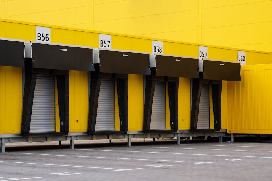 A row of four loading bays attached to a bright yellow warehouse or retail building, each bay labeled B56, B57, B58, and B59 with white signage. The bays feature black retractable shelters and metallic roll-up doors positioned at ground level, with a slightly overcast outdoor setting. The area in front of the bays is paved with parking spaces, some of which are marked with white lines. The image depicts the exterior environment likely used for delivery or removals services, with no vehicles or personnel visible. The clean, organized scene reflects the typical loading area for furniture transport or home relocation logistics, aligning with the services offered by Man with Van Tolworth, who may organize permits for such bays as indicated by the page title.