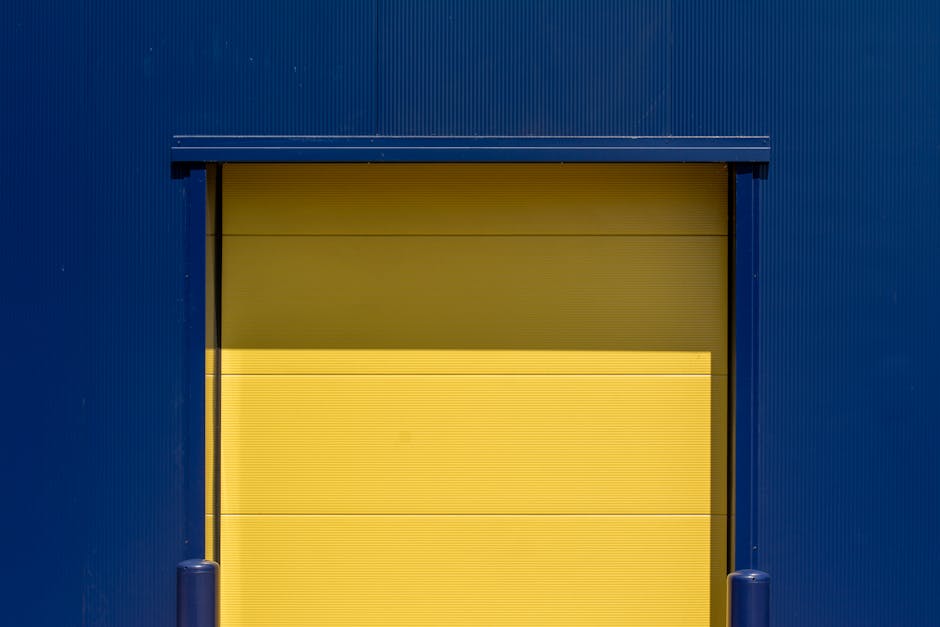A large yellow roll-up loading bay door set within a dark blue industrial building exterior, with visible ridges on the door's surface. The door is partially open, revealing a glimpse of the interior space. On either side of the door, there are blue bollards positioned on the pavement to protect the structure during vehicle loading and unloading. The scene is lit with natural daylight, casting subtle shadows on the door and building façade. This setting captures the typical environment for home relocation and furniture transport operations, where professional removals services like those by Man with Van Tolworth facilitate efficient packing and loading of household items, as teams carefully handle boxes, furniture, and equipment to ensure safe transit.