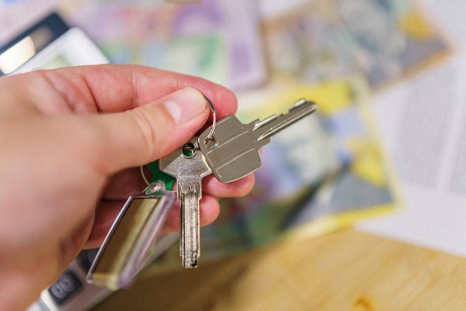 A close-up of a person's left hand holding a set of keys with a metal keyring. The keys include a standard house key and a smaller key, both metallic and shiny. In the background, there are blurred figures of documents, possibly maps or plans, with shades of yellow, purple, and green, suggesting a setting related to home relocation or planning. The person's hand is positioned over a light wooden surface, and the image appears to be taken indoors, potentially in a planning or packing area used by Man with Van Tolworth during a house removal process. This visual can support content about packing, key handover, or planning stages of house removals, aligning with services offered by [COMPANY_NAME].