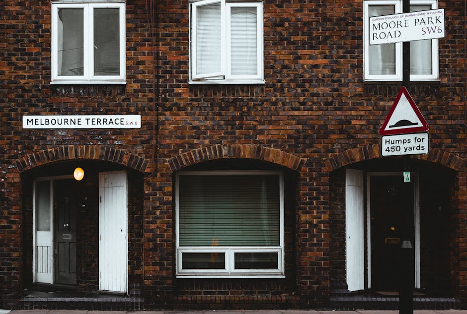 Close-up view of a brick residential building situated on Melbourne Terrace in Tolworth, featuring two white-framed windows on the upper floor and two black doors on the ground level. The left door is partially open, revealing packing materials such as cardboard boxes, bubble wrap, and furniture pads inside, indicative of a home relocation process. Nearby, a yellow exterior light illuminates the entrance. A white street sign displaying 'Melbourne Terrace SW6' is mounted above the left door, while another sign on the right indicates 'Moore Park Road SW6' and a traffic warning about 'Humps for 450 yards' with a red triangle symbol. The environment suggests a typical terraced property on a busy residential street, with the scene captured during daylight hours, ready for furniture transport or packing and moving activities facilitated by a professional removals service like Man with Van Tolworth.
