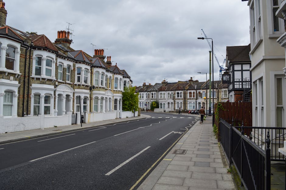 A quiet residential street in Tolworth featuring row houses with Victorian-style architecture, including bay windows, decorative brickwork, and pitched roofs with multiple chimneys. The street has a paved sidewalk on the right side with black metal railings, and a few pedestrians walking along. The road is marked with standard white lines and has parked cars along the curb. The sky is overcast with grey clouds, and a construction crane is visible in the distance, indicating ongoing development. This scene exemplifies an area suitable for home relocation and furniture transport services, with visible urban features like street lighting and traffic bollards. Man with Van Tolworth may assist with packing, moving, and loading furniture and boxes during house removals in this neighbourhood.