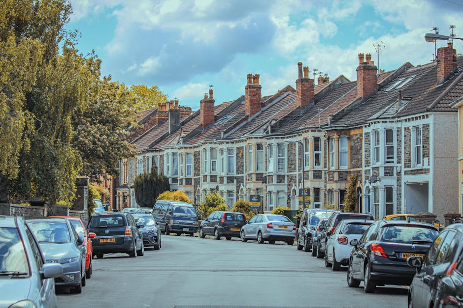 Close-up view of a brick residential building situated on Melbourne Terrace in Tolworth, featuring two white-framed windows on the upper floor and two black doors on the ground level. The left door is partially open, revealing packing materials such as cardboard boxes, bubble wrap, and furniture pads inside, indicative of a home relocation process. Nearby, a yellow exterior light illuminates the entrance. A white street sign displaying 'Melbourne Terrace SW6' is mounted above the left door, while another sign on the right indicates 'Moore Park Road SW6' and a traffic warning about 'Humps for 450 yards' with a red triangle symbol. The environment suggests a typical terraced property on a busy residential street, with the scene captured during daylight hours, ready for furniture transport or packing and moving activities facilitated by a professional removals service like Man with Van Tolworth.
