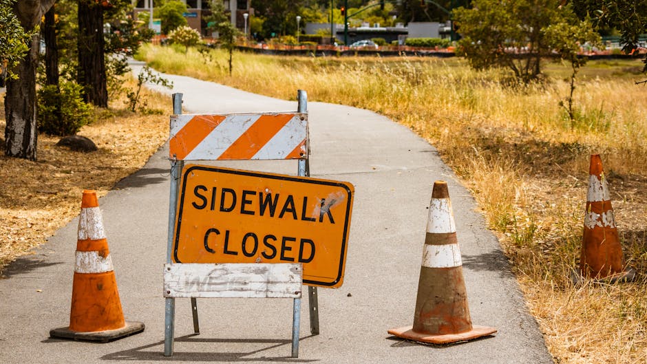 A temporary roadblock setup on a residential sidewalk featuring three orange and white traffic cones and a large rectangular construction barrier with orange and white diagonal stripes. In front of the barrier, a bright orange sign with black text reads 'SIDEWALK CLOSED,' indicating restricted pedestrian access. The sidewalk is narrow, with the closed area leading into a pathway surrounded by dry grass and trees, suggesting an outdoor environment. In the background, there are glimpses of buildings and parked cars, with trees providing partial shade. The scene appears to be daytime, under natural lighting conditions. This setup is indicative of construction, maintenance, or roadworks, which could impact residential moving or delivery schedules. Man with Van Tolworth may need to navigate this temporarily restricted route during home relocation and furniture transport activities, particularly when planning removals connected to the advice on road closure and skip permits for Tolworth moves.