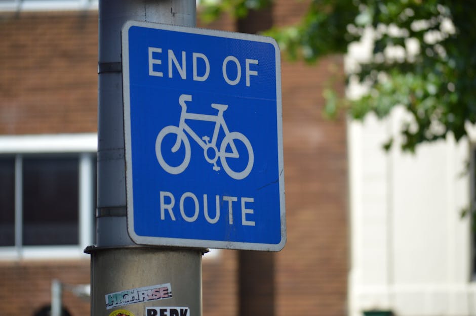 A close-up photograph of a blue and white road sign indicating the 'End of Route' for bicycles, mounted on a grey metal pole outdoors. The sign features a bicycle symbol and text, with a small sticker at the bottom of the pole displaying various colours and text. In the background, there are blurred brick buildings, a window, and some greenery, suggesting an urban environment. This image relates to house removals and moving logistics, emphasizing the importance of understanding local traffic and route restrictions during home relocations, which services like those provided by Man with Van Tolworth assist with in planning and executing smooth furniture transport and packing and moving activities.