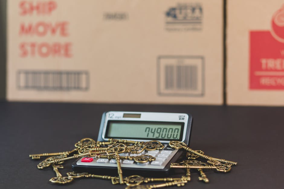 A close-up of a calculator displaying the number 749800 placed on a black surface, surrounded by numerous small brass keys and keychains strewn across the top. In the background, there are several large cardboard boxes with printed labels and barcodes, indicating they are packed for moving or storage. The boxes are positioned against a blurred background, suggesting an indoor setting used for packing or home relocation. The scene illustrates elements involved in the logistics of house removals, such as packing materials, and highlights the detailed process of moving, which may include valuation, organization, and transportation of household items. Man with Van Tolworth, a professional removals service, often handles these activities as part of their same-day home removal operations in Tolworth.
