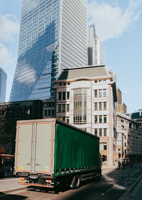A large green curtain-sided delivery truck is parked on a city street in front of a row of modern glass skyscrapers and older stone buildings, with several pedestrians walking nearby. The truck is positioned close to the sidewalk, with its rear doors closed and secured, ready for furniture transport or packing and moving activities as part of a home relocation process. The urban environment is characterized by high-rise office buildings reflecting the sky, contrasting with the older architecture at street level. The truck is situated on the roadway, and the area appears to be a busy commercial district during daylight hours, with clear weather and bright natural light illuminating the scene. This image illustrates the typical scene of loading or unloading during professional house removals and moving services provided by companies like Man with Van Tolworth, supporting efficient furniture transport and logistical operations in urban environments.