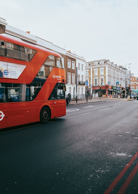 A red double-decker bus traveling along a street in Tolworth, with residential and commercial buildings lining the pavement. The buildings are a mix of brick and stucco facades, some with shopfronts and signage visible. The street is relatively quiet with a few pedestrians and parked vehicles. In the foreground, the lower part of the bus and part of the road markings can be seen, indicating a typical urban environment suitable for home relocations or moving services. The scene is well-lit with natural daylight, providing a clear view of the surrounding architecture and street layout. This image aligns with the theme of house removals and furniture transport, reflecting the busy, accessible urban setting where [COMPANY_NAME] operates, particularly in the context of planning routes and parking tips for smooth removals in Tolworth.