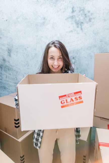 A woman with long brown hair, smiling at the camera, is holding an empty cardboard box labeled 'GLASS' in bold red letters, with protective foam padding visible inside. She is standing inside a room with a light grey textured wall behind her, surrounded by several other packed cardboard boxes of various sizes, some sealed with red or black tape. The boxes are stacked on the floor and appear to be prepared for a home relocation or furniture transport. The scene is well-lit, indicating a clean and organized environment typical of packing and moving processes, supported by the services of Man with Van Tolworth in relation to home removals and moving logistics.