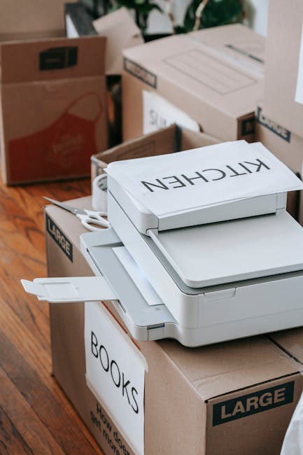 A close-up image showing several cardboard boxes and packaged office equipment on a wooden floor inside a property. In the foreground, there is a white multifunction printer or scanner with a label ma