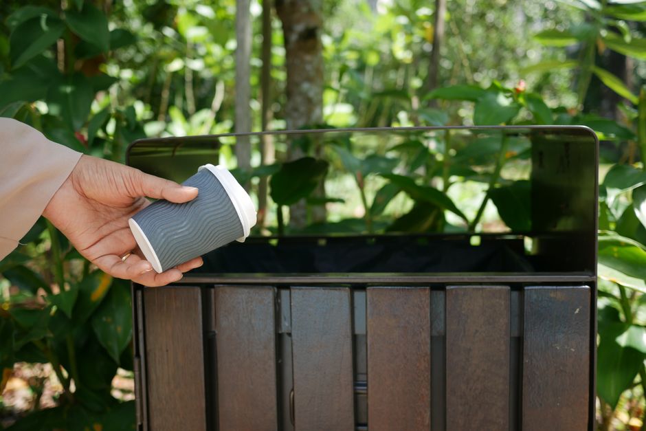 A person holding a grey paper coffee cup with a white lid is placing it into a large, open, black metal container situated outdoors among lush green foliage and plants. The container appears to be part of a home relocation or packing process, as it is positioned on a paved surface next to a wooden structure or fence, with natural daylight illuminating the scene. The image depicts the careful handling involved in packing and transporting items during a house move, with the container possibly used for waste disposal or to organize belongings before removal. The background includes various leaves and stems, emphasizing an outdoor setting, and the container is open, showing space for additional items or waste to be collected, aligning with logistics for furniture transport or packing during moving services provided by Man with Van Tolworth.