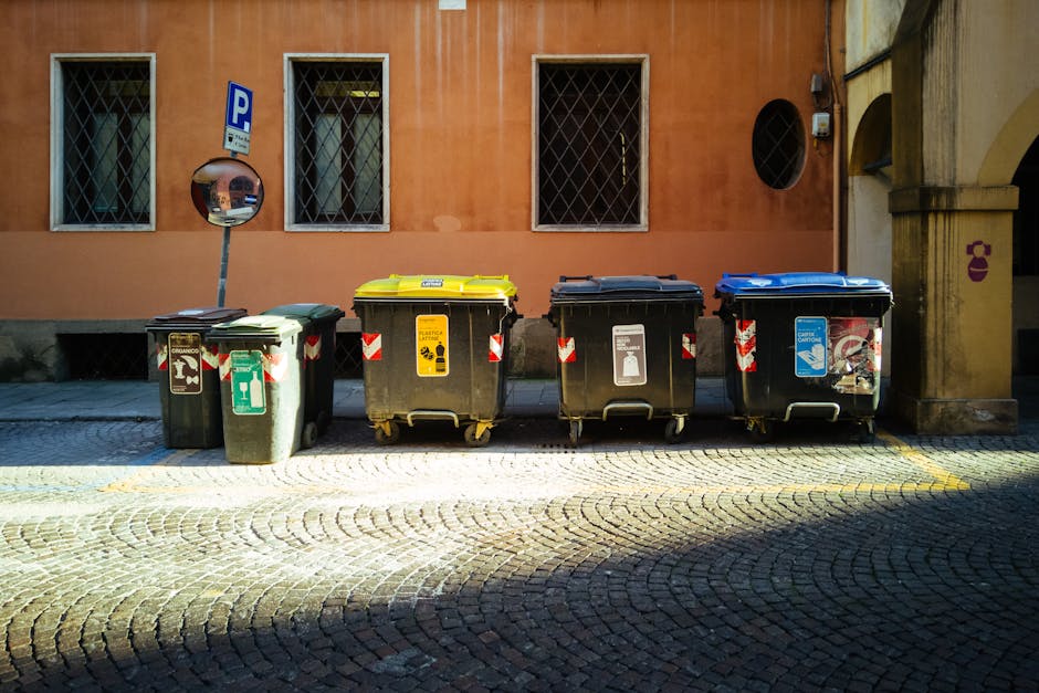 An outdoor scene on a cobblestone street in Tolworth showing five wheeled recycling bins lined up against a pinkish-orange building wall. The bins are color-coded with green, yellow, black, black, and blue lids, each designated for different waste or recycling types, with visible labels and symbols. The green bin has a recycling symbol and a green sticker, the yellow bin is marked for packaging waste, the next black bin has a label for general waste, and the blue bin appears for paper or cardboard. A curved bollard with a mirror is positioned behind the green bin, reflecting part of the street. The building features rectangular windows with iron grills and a small circular window, and there is an electric meter or box attached to the wall near the corner. The scene suggests a typical urban waste disposal area, relevant to residential or home relocation services by Man with Van Tolworth, with an environment suitable for loading or unloading relevant to furniture transport and packing processes.
